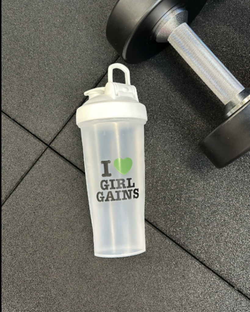 Clear protein shaker with 'I ❤️ Girl Gains' text on a gray floor with a dumbbell.